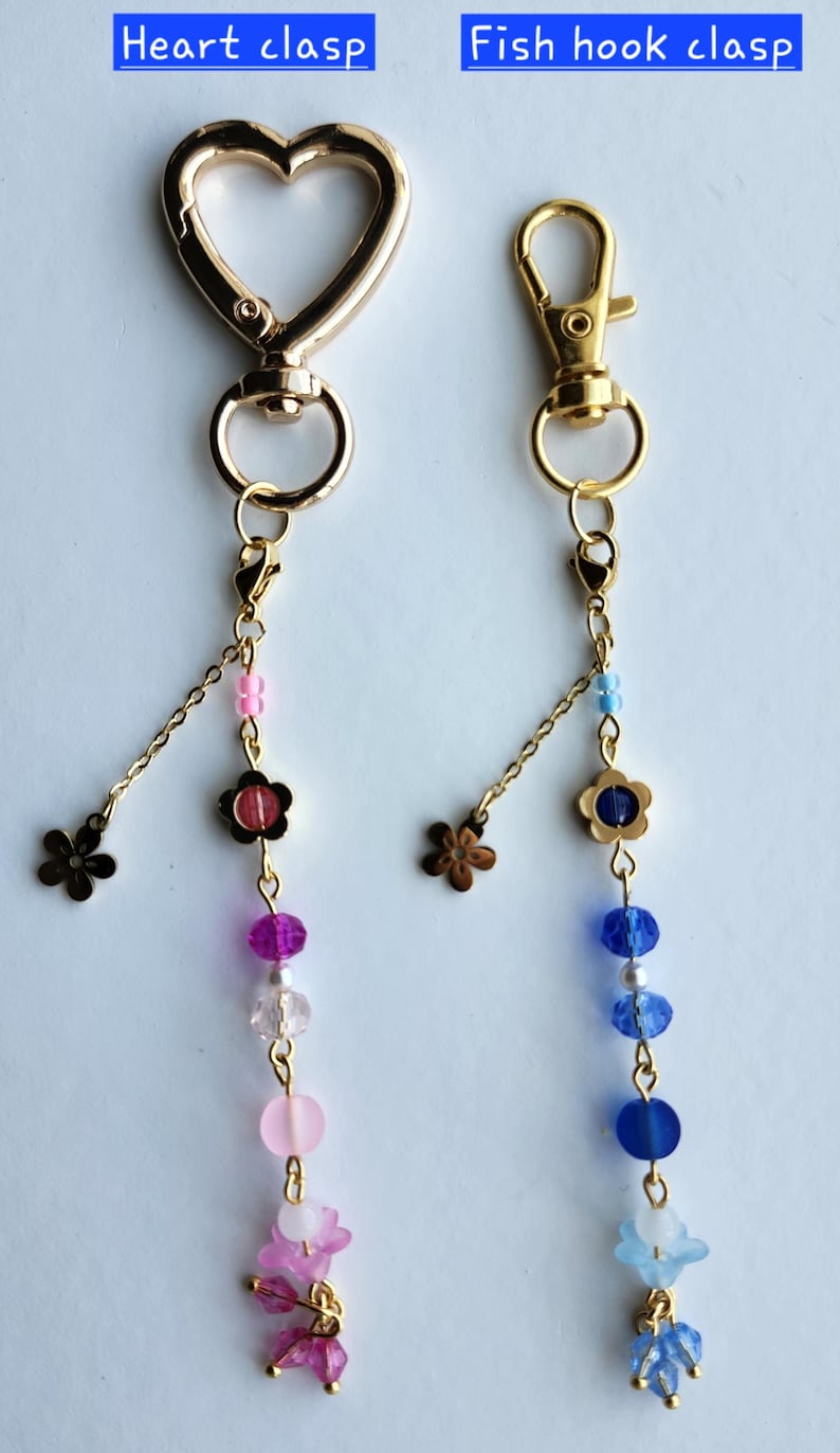 May include: Two gold keychains with different clasps. The keychain on the left has a heart clasp and a pink and purple beaded charm. The keychain on the right has a fish hook clasp and a blue and white beaded charm. Both charms have a small flower charm at the top.