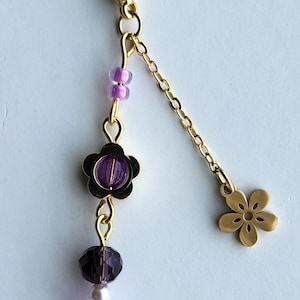 May include: A gold keychain with a purple and white beaded charm. The charm has a flower design and a small gold flower charm at the end of the chain.