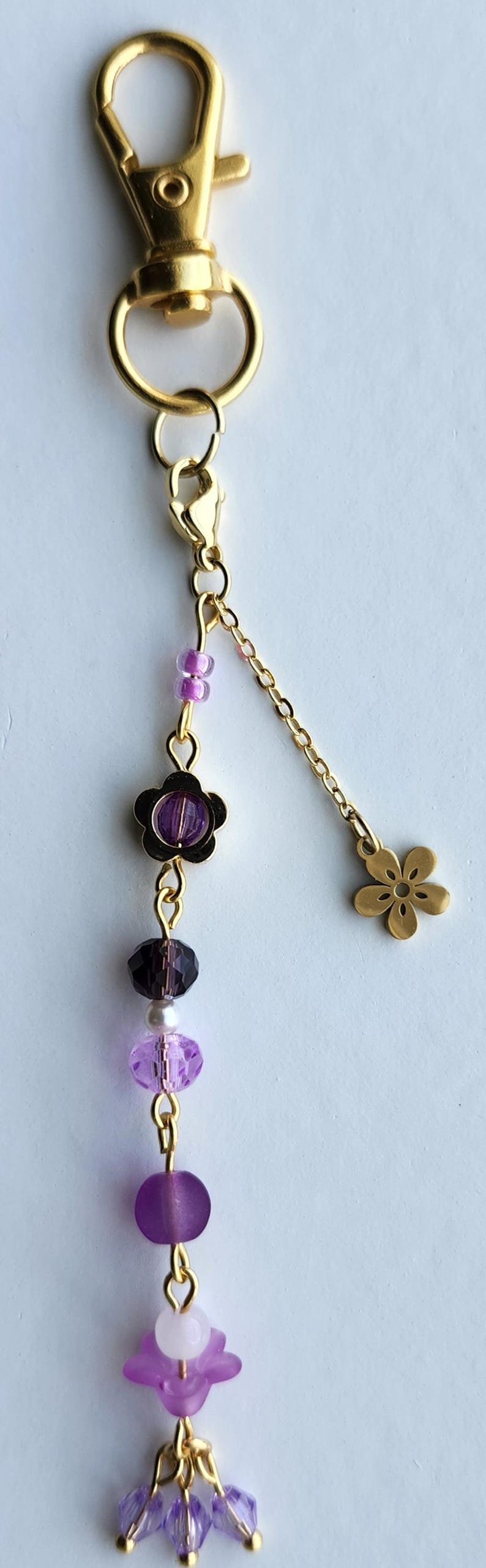 May include: A gold keychain with a purple and white beaded charm. The charm has a flower design and a small gold flower charm at the end of the chain.