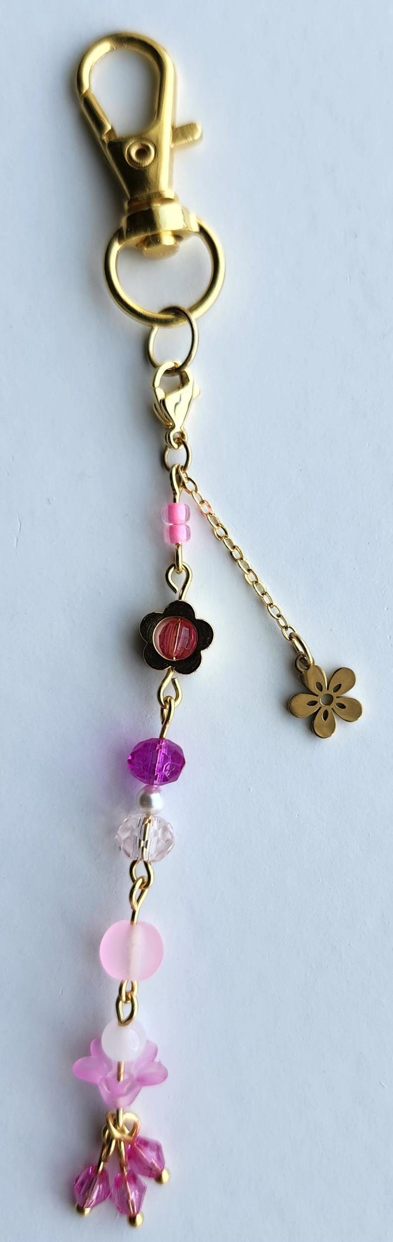 May include: A gold key chain with a pink and purple flower charm. The charm has a pink flower with a white center and a purple flower with a clear center. The key chain also has a small gold flower charm.