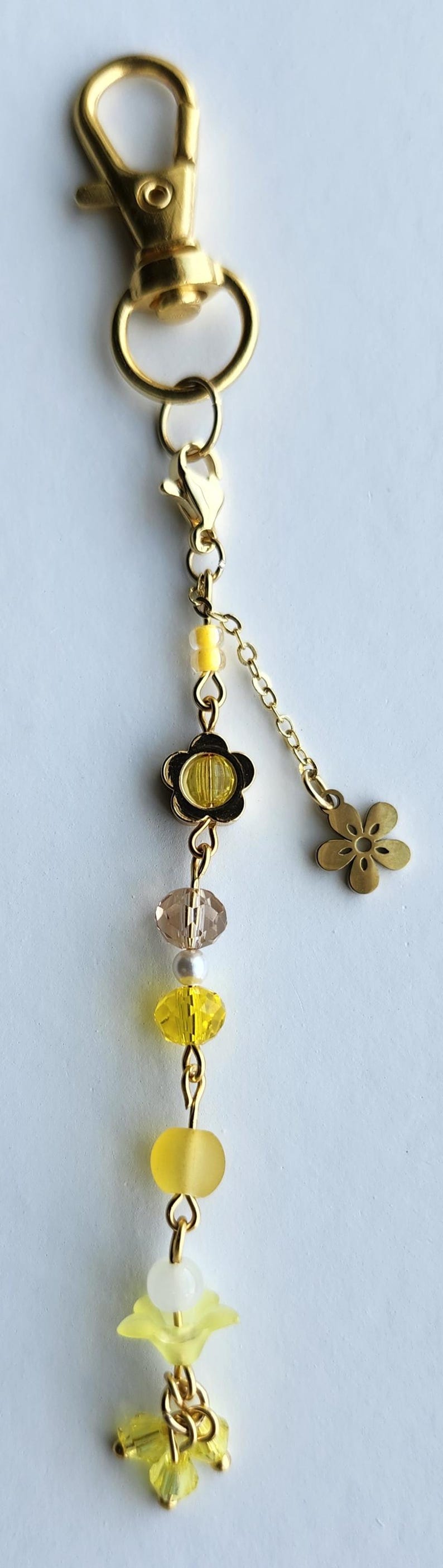 May include: A gold keychain with a yellow flower charm, yellow and clear glass beads, and a small gold flower charm. The keychain is attached to a gold split ring.