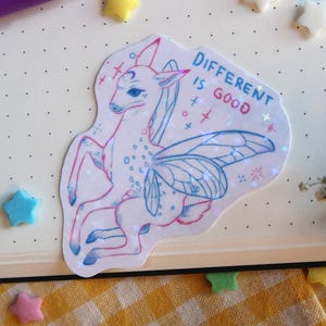 Könnte beinhalten: Ein holografischer Aufkleber mit einem fabelhaften Wesen mit hirschähnlichen Merkmalen und Schmetterlingsflügeln. Der Aufkleber trägt den Text "DIFFERENT IS GOOD". Sterne und rosa und lila Stifte umgeben das Bild.