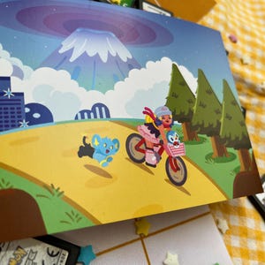 Może przedstawiać: Kolorowa ilustracja przedstawia osobę na r&oacute;żowym rowerze z koszykiem, w towarzystwie niebieskiego stworzenia. Tło przedstawia g&oacute;rę, drzewa i panoramę miasta. Widoczne są r&oacute;wnież kartridże do gier i dekoracje w kształcie gwiazd.