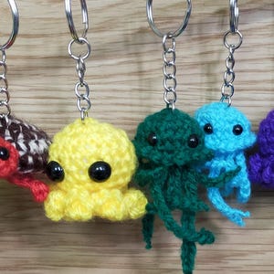 May include: A selection of handmade crochet keyrings depicting sea creatures. The keyrings include a red octopus, a turtle, a yellow octopus, green and blue jellyfish, a purple octopus, and a white jellyfish. Each keyring has a silver chain and ring.