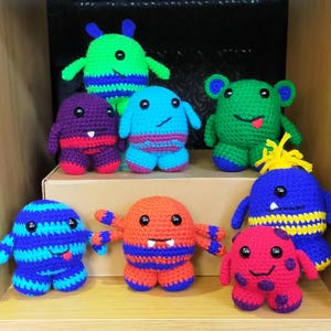 May include: A collection of eight crocheted stuffed toys in various colours and designs, including a green monster, a blue monster, a purple monster, a red monster, a yellow monster, a striped monster, a spotted monster, and a monster with a yellow mohawk.