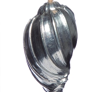 May include: A silver-coloured, decorative pull with a spiral design, suspended by a natural-coloured rope. The pull has a bulbous shape with vertical grooves, and is likely used for a drawer or cabinet.