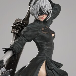 Peut inclure: Figurine détaillée d'un personnage du jeu vidéo Nier: Automata. La figurine porte une robe noire fendue, un bandeau et des cheveux courts blancs. Elle tient une grande arme sombre. Le texte "TANUKI FIGURES" est visible.