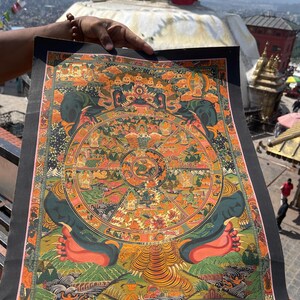 May include: A colorful, detailed Buddhist Thangka painting depicting the Wheel of Life. The artwork features a central circular design with various figures and scenes, framed by a black border. The painting is held up against a backdrop of a golden stupa and a cityscape.