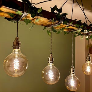 Könnte beinhalten: Ein rustikaler Holz-Kronleuchter mit fünf Edison-Glühbirnen, die daran hängen. Das Holz ist verwittert und hat eine natürliche Oberfläche. Die Glühbirnen sind aus klarem Glas und strahlen ein warmes Licht aus. Der Kronleuchter ist mit grünen Efeu-Ranken verziert.