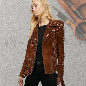 May include: A brown suede jacket adorned with gold studs. The jacket has an asymmetrical zip, notched lapels, and a belted waist. The sleeves feature zips and stud detailing. The jacket is worn over a black top and black trousers.
