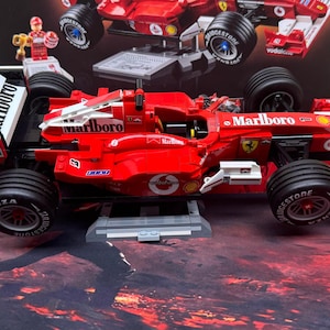 May include: A red and white Formula 1 race car model with black tires. The model features the "Marlboro" logo and other sponsor decals. A small figure is visible on the top left. The car is displayed on a gray base.