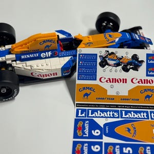 May include: A blue, white, and orange Lego race car model with the number 5. The car features sponsor logos including "Canon", "Camel", and "Labatt's". A sheet of matching stickers is next to the car.