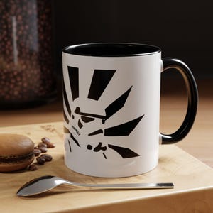 May include: A white ceramic mug with a black rim and handle. The mug features a black and white graphic of a Star Wars Stormtrooper helmet with a sunburst design behind it.