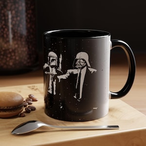 Könnte beinhalten: Schwarze Kaffeetasse mit einer weißen Grafik von Darth Vader und Boba Fett in einer Szene, die an den Film Pulp Fiction erinnert. Die Tasse hat einen schwarzen Henkel und ein schwarzes Innenleben.