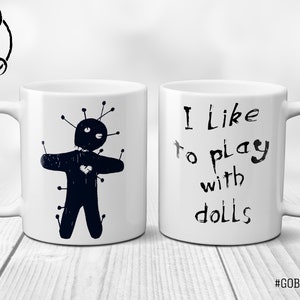 May include: White ceramic mug with a black voodoo doll illustration on one side and the text "I like to play with dolls" on the other side. The text "#GOBLINDREAMS" is printed on the bottom of the mug.