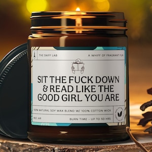 Op de afbeelding: Een bruine glazen potkaars met een zwart deksel, op een houten oppervlak. Het etiket luidt "SIT THE FUCK DOWN & READ LIKE THE GOOD GIRL YOU ARE". De kaars is gemaakt van natuurlijke sojawas en een katoenen lont, met een brandtijd van maximaal 50 uur.