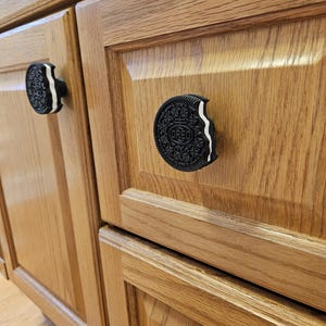 May include: Close-up of wooden cabinet doors with Oreo cookie-shaped cabinet knobs. The knobs are black with white cream filling and the Oreo logo. The cabinet doors are a light brown color.