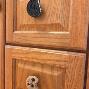 May include: Two cabinet knobs shaped like cookies. The top knob is a black and white sandwich cookie with a bite taken out, featuring the text OREO. The bottom knob is a tan chocolate chip cookie with dark brown chips and a bite taken out.