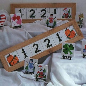 May include: A set of hand-painted ceramic tiles with various designs, including numbers, hearts, pumpkins, snowmen, and a shamrock. The tiles are displayed on small wooden stands.