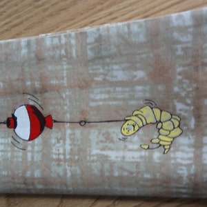 May include: A beige fabric pouch with a red and white bobber and a yellow worm on a fishing line printed on it.