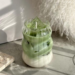 May include: A green and white layered candle in a clear glass jar with a wavy design. The candle has a white wick and is surrounded by ice cubes.