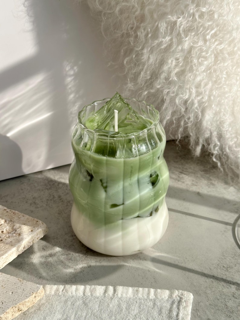 May include: A green and white layered candle in a clear glass jar with a wavy design. The candle has a white wick and is surrounded by ice cubes.