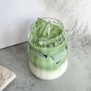 May include: A green and white candle in a glass jar shaped like a cup of iced matcha. The candle has a white wick and is surrounded by clear ice cubes.