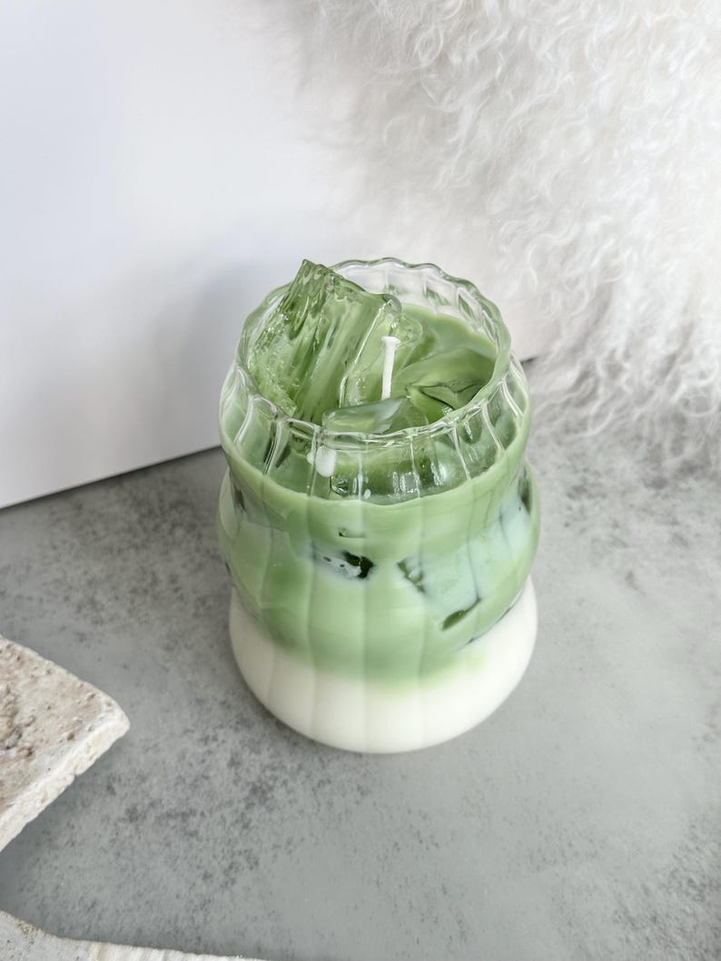 May include: A green and white candle in a glass jar shaped like a cup of iced matcha. The candle has a white wick and is surrounded by clear ice cubes.