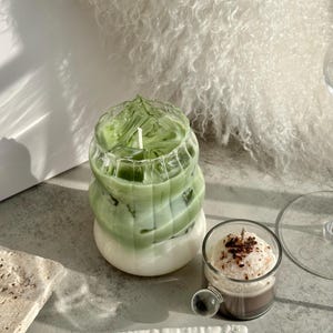 May include: Two glass candle jars with a green and white layered candle inside. The candle on the left is a green and white layered candle. The candle on the right is a brown and white layered candle.