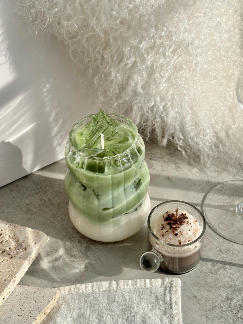 May include: Two glass candle jars with a green and white layered candle inside. The candle on the left is a green and white layered candle. The candle on the right is a brown and white layered candle.
