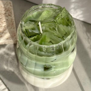 May include: A clear glass candle jar with a green and white layered candle. The candle is shaped like a drink with ice cubes and a white wick.