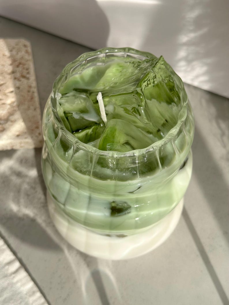 May include: A clear glass candle jar with a green and white layered candle. The candle is shaped like a drink with ice cubes and a white wick.