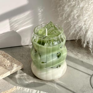 May include: A green and white layered candle in a clear glass jar with a textured surface. The candle has a white wick.