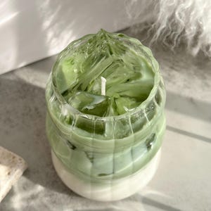 May include: A green and white candle in a glass jar shaped like a cup of iced matcha. The candle has a textured surface and a white wick.