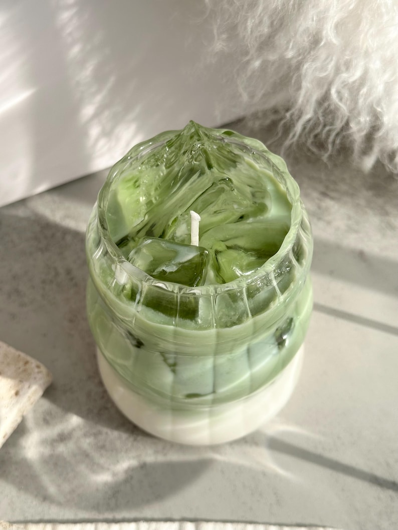 May include: A green and white candle in a glass jar shaped like a cup of iced matcha. The candle has a textured surface and a white wick.