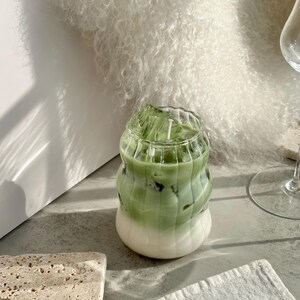 May include: A green and white iced drink in a clear glass with a wavy design. The drink is sitting on a light gray surface.