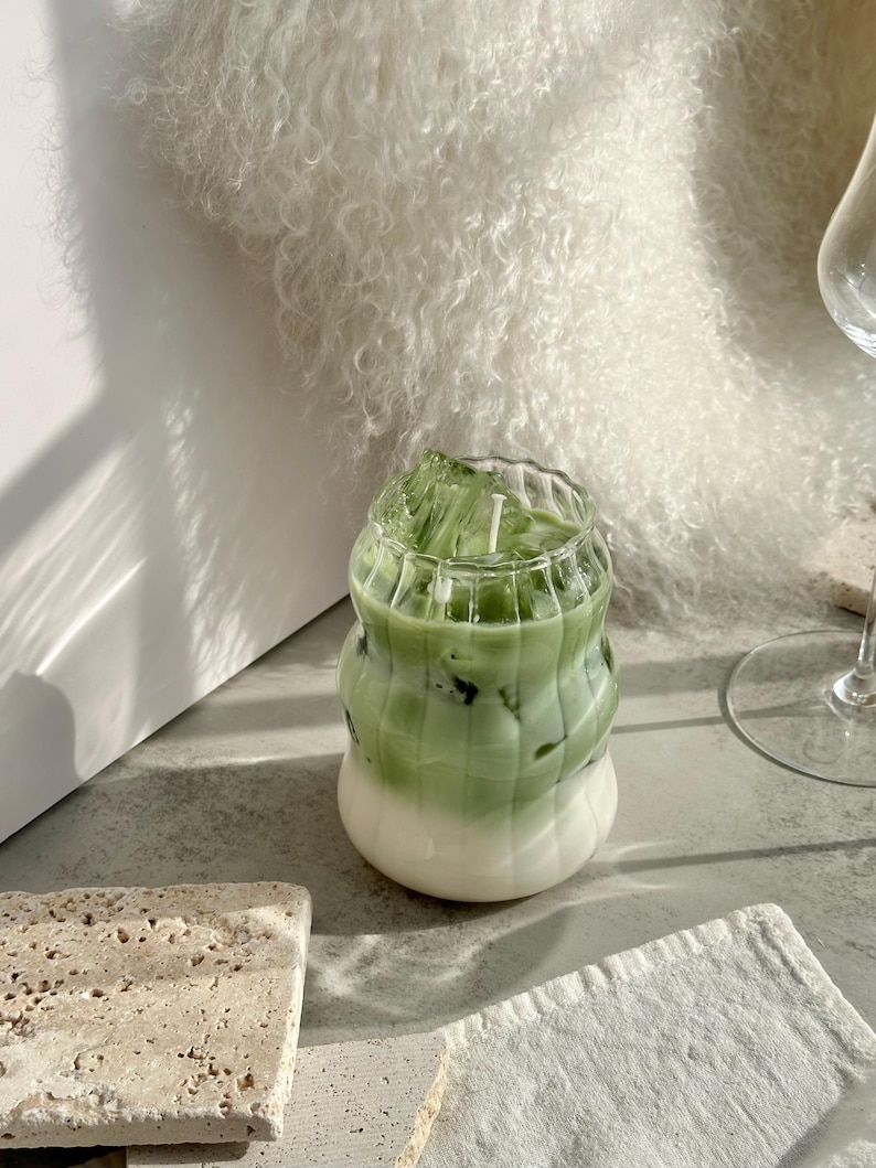 May include: A green and white iced drink in a clear glass with a wavy design. The drink is sitting on a light gray surface.