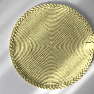Può includere: Un sottobicchiere rotondo giallo chiaro con un bordo intrecciato. Il centro presenta un motivo a spirale. Il sottobicchiere è realizzato in materiale tessuto. Una piccola etichetta rettangolare è attaccata al bordo.
