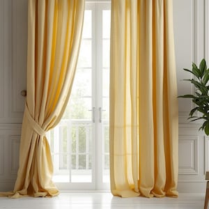 May include: Pale yellow linen curtains hang in front of a white door and window. The curtains are tied back on one side, revealing a glimpse of the outdoors. A potted plant and a beige armchair are in the background, creating a bright and airy interior design.