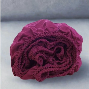 May include: A rolled-up, deep burgundy fitted sheet. The sheet is made of a soft fabric and has elasticated edges. The fabric is bunched up, showing the texture of the material. The sheet is designed for a bed.