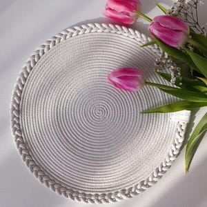 May include: A round, off-white woven placemat with a braided edge. The placemat is decorated with three pink tulips and small white flowers with green leaves. The placemat is approximately 30 cm in diameter.