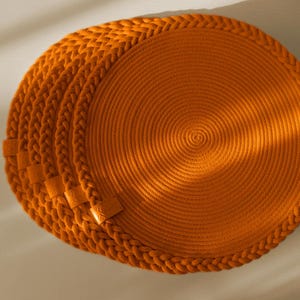 Peut inclure: Un ensemble de sets de table ronds d'un orange vif, fabriqués à partir de corde enroulée. Chaque set présente une bordure en corde tressée et une petite étiquette rectangulaire en similicuir fixée sur le bord, avec un motif circulaire.