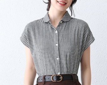 May include: A black and white gingham short-sleeved shirt with a collar and button closure. The shirt is worn with a brown skirt and a black belt with a gold buckle.