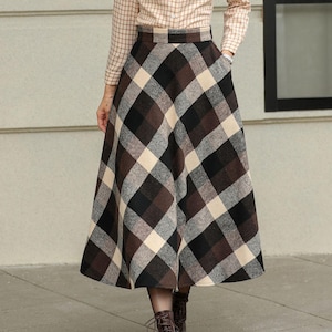 May include: A woman wearing a beige plaid button-down shirt and a long, brown, black, and cream plaid skirt. The outfit is paired with brown boots. The skirt has a high waist and a flared silhouette.