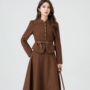 May include: A brown skirt suit with a fitted jacket and a flared skirt. The jacket has a mandarin collar and gold buttons. The skirt has a brown leather belt with a gold buckle. The model is wearing brown lace-up boots.