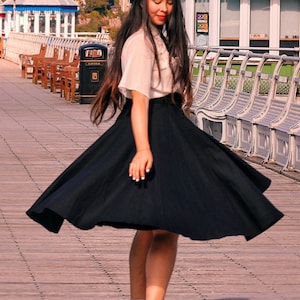 Peut inclure: Une femme portant une blouse blanche, un b&eacute;ret noir et une jupe noire se tient sur une jet&eacute;e en bois. Sa jupe flotte au vent. Elle porte des bottes noires et a de longs cheveux noirs.