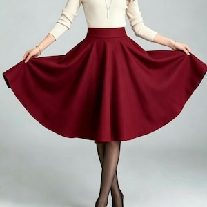 May include: A cream-colored long-sleeve mock neck top paired with a solid deep red high-waisted midi skirt. The skirt features a wide waistband and a full, flared silhouette that extends outward. The outfit is completed with sheer black tights.