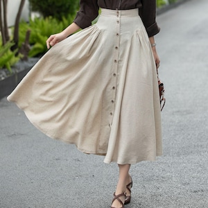 Puede incluir: Una mujer con una camisa marrón oscuro y una falda midi beige abotonada. Lleva zapatos marrones. El fondo incluye follaje verde y un camino pavimentado. La falda tiene un diseño abotonado.