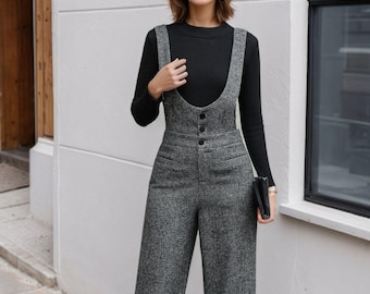 May include: A woman wearing a black long-sleeve top and gray tweed overalls with wide legs. The overalls have a bib front with three buttons and adjustable straps. She is also wearing black boots and holding a black clutch.