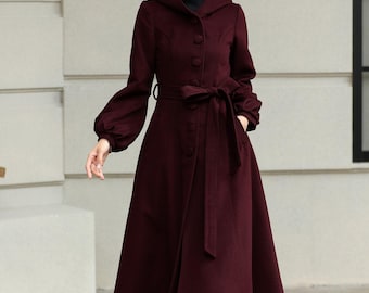 May include: A burgundy hooded coat with a button-down front and a matching belt tied at the waist. The coat features long sleeves with a slight puff at the shoulders and a flared skirt. The person is wearing black boots.